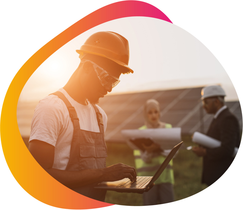 profile of a man with glasses and a hard hat on with computer and two people standing in the background with site maps in front of a solar panel