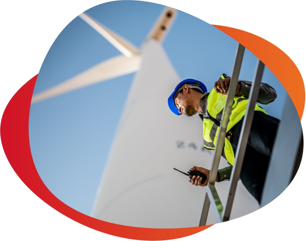 operations person wearing a vest, hard hat, and a walkie talkie in hand below a wind turbine